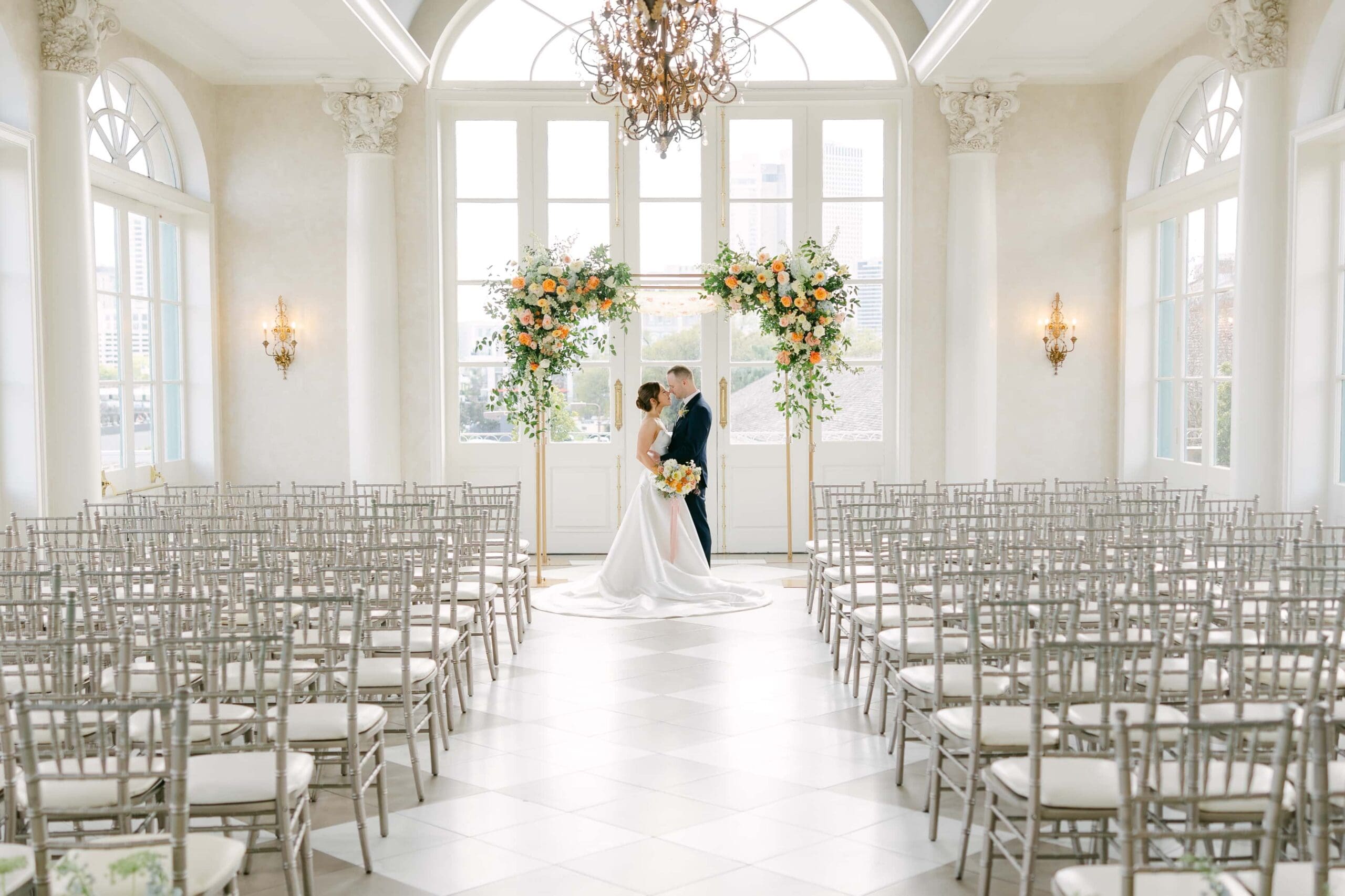 Bride and groom dressed in wedding attire standing under colorful floral chuppah in Marche, a French quarter wedding venue. Couple is standing in a white room with multiple large domed windows.