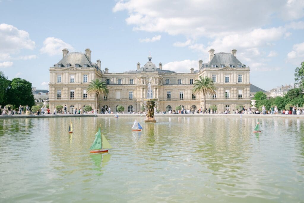 French building with a blue, cloud filled sky above. A fountain with little toy sail boats in the foreground.
