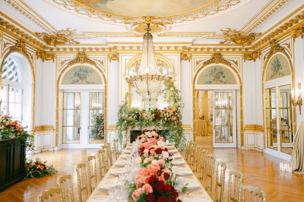 Image of a wedding reception table scape with red and pink flowers as the centerpiece. Room is decorated with a grand chandelier, gold accents, and a celestial mural painted on the ceiling.